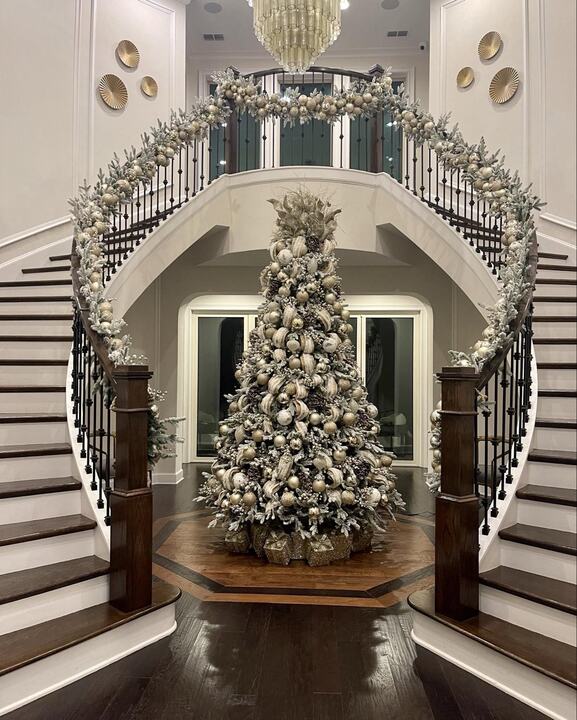 A tree centered in a grand foyer between two curved staircases dressed in garland